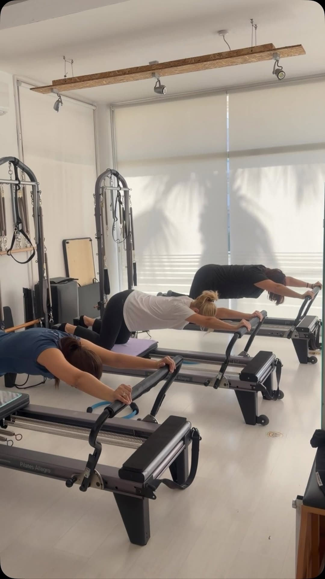Pilates studio group class with three people performing stretching exercises on reformer machines under soft natural light