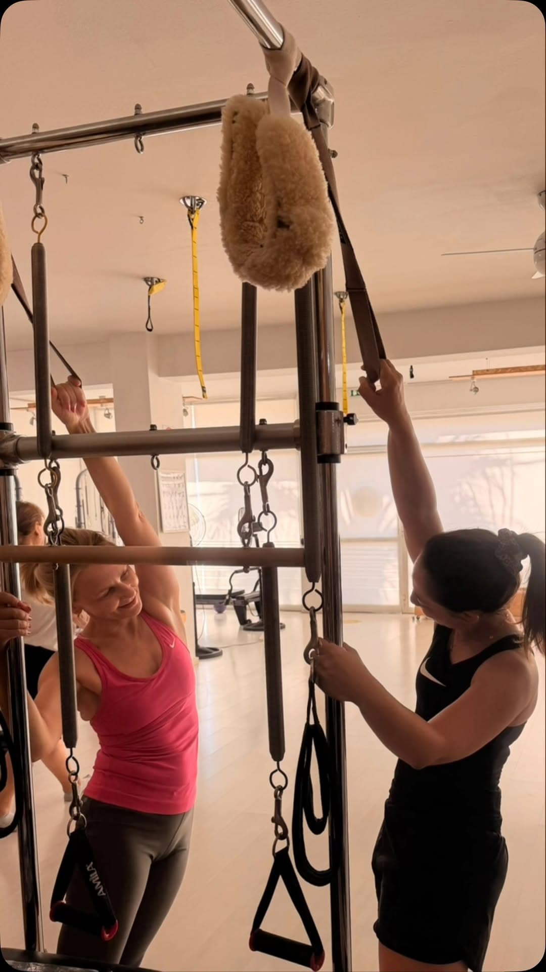 Pilates studio workout action with two women exercising using Pilates equipment and straps, one wearing a pink top and the other in black, in a bright indoor fitness studio