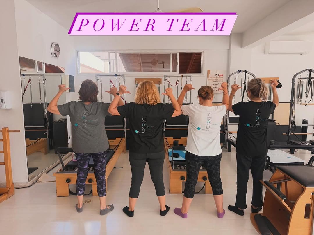Four women standing with backs to the camera inside a pilates studio, pointing thumbs at the words 'Power Team' above their heads
